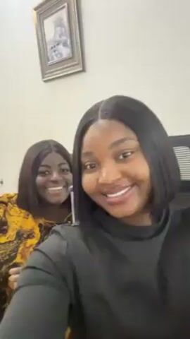 Two women, one in a black long-sleeved shirt and another in a yellow and black patterned top, are in an indoor setting, possibly an office, as suggested by the office chair visible. The woman in black is in the foreground, making a funny face at the camera. The other woman is smiling in the background. A framed picture hangs on the light-colored wall.