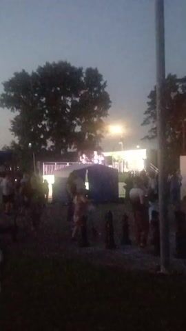 The video shows a nighttime outdoor gathering. A stage with lights and people on it is visible in the background, potentially a performance or event. In the foreground, there are various people milling around. Scattered on the ground are large chess pieces, suggesting a giant outdoor chess set. Trees and a street light are also present in the environment. The overall atmosphere appears to be festive and social.