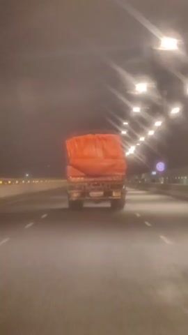 The video shows the rear view of a truck driving on a highway at night. The truck is covered with an orange tarp. Streetlights illuminate the highway, and the truck's taillights are visible. The road is wet, and there are white lane markings. The video is shaky, suggesting it was filmed from a moving vehicle.