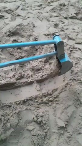 The video shows a close-up view of a person's hands using a tool with a blue handle and a metal digging end to move sand. The tool is being used to dig into the sand and displace it. The environment appears to be a sandy area, possibly a beach or sandbox, with footprints visible in the sand. The video is shot from a low angle, focusing on the action of digging.