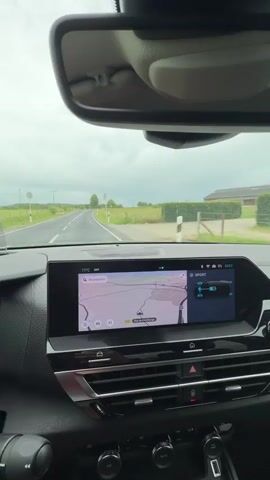 The video captures a first-person perspective from inside a car driving along a rural road. The car's dashboard and the view through the front windshield are prominent.

The infotainment screen in the center of the dashboard displays a navigation map on its left side, showing roads like "Rue de Luxembourg," "Rue de Schifflange," and the "A4" highway, consistent with a location in Luxembourg. The map includes a 3D view option and an alert icon. On the right side of the screen, a car settings interface indicates "SPORT" mode is selected, accompanied by a graphic possibly showing battery or power usage. Other details on the screen include a temperature reading of "17°C" and the time "13:57." Various physical buttons and control knobs are visible below the screen.

Through the windshield, a two-lane asphalt road can be seen, gently curving to the right. Green fields and grass stretch out on both sides of the road, with scattered trees and hedges. A barn or agricultural building is visible in the distance on the right. The sky is overcast with gray clouds, and small raindrops are visible on the windshield, suggesting recent or ongoing light rain. Briefly, a red and white triangular warning sign for a double bend appears on the right side of the road.

The activity is driving through the countryside on a cloudy afternoon.