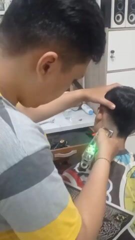 The video shows a person in a gray and yellow striped shirt using an electric hair clipper with a green light to cut the hair on the back of another person's head. The person getting their hair cut is wearing a cape with a colorful comic book design and is seated. In the background, there is a white cabinet with speakers on top, a shelf with scissors, a spray bottle, and various barber tools. A mirror reflects the scene, showing both individuals and the surrounding environment. The person in the striped shirt is holding the head of the person being cut steady with their left hand while operating the clippers with their right. The action is focused on the nape of the neck.