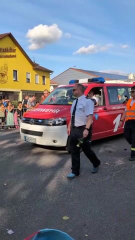 A red fire engine with blue lights and the number 1 on its side is parked on a street. A man in a white shirt and tie walks in front of the fire engine. A person in an orange vest walks beside the fire engine, and another person in an orange vest walks behind the fire engine, pulling a red trailer with the words "JUGENDFEUERWEHR" on it. Several people are standing on the sidewalk in the background. A yellow building with a sign that says "dtstädter Bäckerei" is visible on the left side of the frame. The sky is blue with some white clouds.