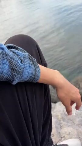 The video shows a person sitting on a rocky surface next to a body of water. The person is wearing black pants and a blue and black plaid long-sleeved shirt. The person's hand rests on their knee. The camera then pans up to reveal the water, which is clear and slightly rippled, with some rocks visible near the shore. The opposite shore is visible in the distance, with buildings and green vegetation covering the hillside. The sky is partly cloudy with white clouds against a blue background.
