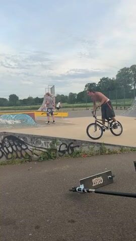 A man is riding a black BMX bike at a skatepark. He is shirtless and wearing dark shorts, white socks, and white Adidas shoes. The bike has black tires, and the handlebars are slightly raised. The man rides across a low concrete barrier covered in colorful graffiti, then continues towards the center of the skatepark, where several ramps and bowls are located. Another person, wearing a black shirt and jeans, is riding a scooter in the background. The skatepark is surrounded by grass, trees, and a chain-link fence. The sky is overcast, and several light poles are visible in the distance.