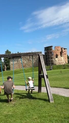 A man and a child are on a playground with a swing set in front of a castle ruin. The man is sitting on a swing on the left, wearing a t-shirt and shorts. The child is sitting on a swing on the right, wearing a white t-shirt, white shorts, and a white hat. The child is swinging forward and backward. The castle ruin is visible in the background, with scaffolding on the right side. The sky is blue with some clouds. The ground is covered in green grass.