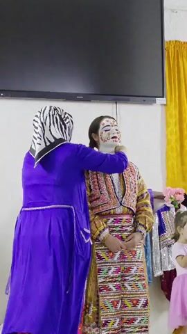 A woman in a blue robe and headscarf with zebra stripes is adjusting the braids of another woman who is wearing a white base makeup with floral decorations on her face and a brightly colored, ornate traditional outfit. The woman with the floral makeup is looking straight ahead. In the background, there are other traditional outfits hanging on a rack, and a small child in a pink dress is visible on the right. The environment appears to be indoors, possibly a backstage area or a preparation room for a cultural event.