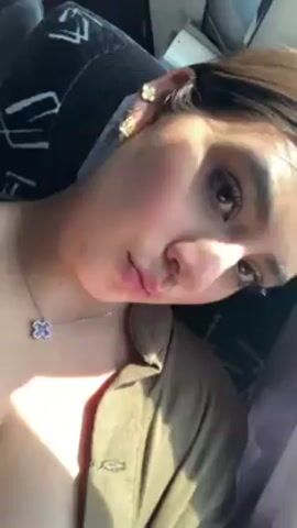The video shows a woman in a vehicle. She is wearing a necklace, earrings, and a jacket. The window shows trees and other vegetation passing by. A seatbelt is visible. There is audio of a man speaking.