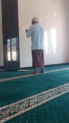 A man wearing a light blue shirt, a white cap, and a brown sarong is shown standing on a green carpet with a patterned border. He is facing away from the camera, towards a wall with a dark vertical panel on the left and windows on the right. He then performs a bowing motion, followed by kneeling on the carpet. There is a small prayer mat visible on the carpet near him. The room appears to be indoors, possibly a mosque, with natural light coming from the windows.
