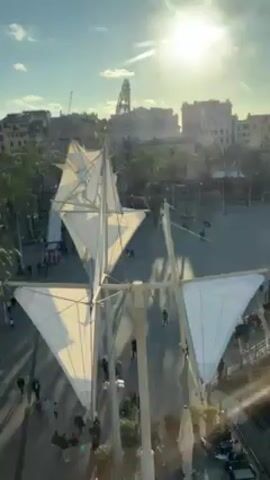 The video shows a view of a public square in what appears to be an Italian city. The view is taken from an elevated position. At the center of the frame is a modern sculpture made up of white triangular sails mounted on poles. Below this are palm trees that line a pedestrian walkway. People can be seen walking along the walkway. In the background are buildings of various heights and colors. A construction crane is visible in the distance. The sun is shining brightly in a blue sky. The video appears to be shot from behind a window.