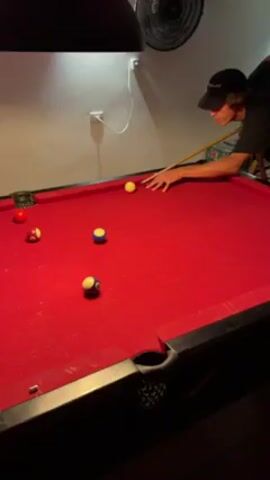 A young man wearing a baseball cap is lining up a shot on a red pool table.  There are several pool balls on the table, including red, yellow, and blue striped balls. A lamp hangs over the pool table, and a fan is visible on the wall behind him. He is holding a pool cue and appears focused on the shot.