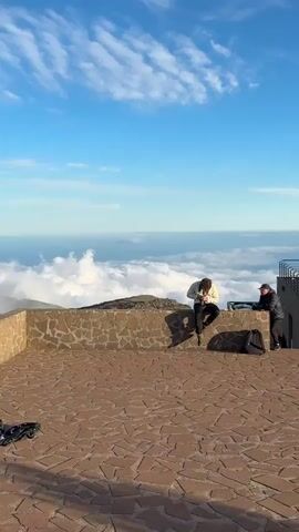 A low-angle shot shows a man sitting on a low stone wall. He is wearing a light-colored top, dark pants, and dark boots. He has curly hair and is holding a camera, looking down at it. Behind him and below the wall, a sea of white clouds stretches into the horizon. A rocky outcropping is visible in the middle of the clouds. The sky is a clear blue with some wispy clouds near the horizon. To the right of the man, another person stands with a backpack nearby. The area in front of the wall is paved with stones. The light suggests it is either early morning or late afternoon.