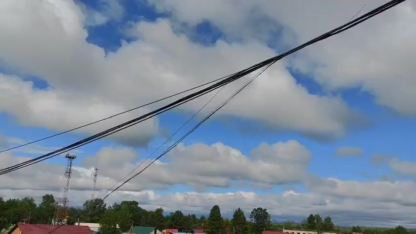 This is a wide shot video of a rural village on a cloudy day. The foreground is dominated by thick black wires strung across the sky, obscuring the view of the village below. In the background, a collection of small houses with red and blue roofs can be seen, interspersed with trees. A tall telecommunication tower is visible to the left of the frame. The sky is a mix of blue and white clouds, with the sun appearing to be behind the clouds. The overall impression is of a somewhat isolated and undeveloped area.