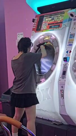 A person wearing a grey off-the-shoulder t-shirt, a black skirt, and black gloves plays an arcade game. The game machine is white with a large circular screen that displays colorful patterns and game graphics. The player is actively touching the screen with their gloved hands, indicating they are interacting with the game. The background is a pink wall with some posters. To the left of the game machine is a black trash bin. To the right of the game machine, part of another similar arcade machine is visible. The overall environment appears to be an arcade or entertainment venue.