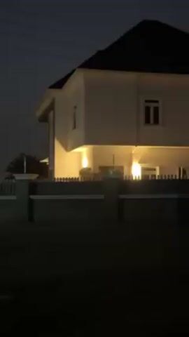 The video shows the exterior of a two-story house at night. The house is painted white and has a dark roof. It's surrounded by a dark gray fence with pillars. Yellow lights illuminate the area under the overhang of the second story. There's a dark grassy area in front of the fence. Power lines are visible in the dark sky above.