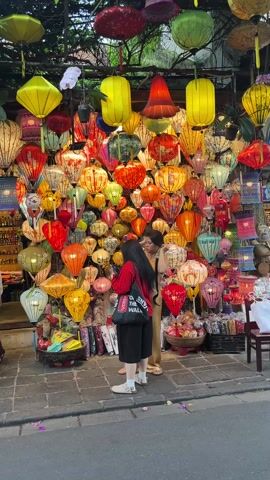 The video shows two women standing in front of a shop displaying a vibrant collection of lanterns. The lanterns, hung closely together, create a colorful wall of different shapes and sizes in shades of red, orange, yellow, and green. The women appear to be looking at the lanterns. One woman is wearing a red shirt and black pants with a black tote bag, while the other woman is wearing a tan dress. The shop also displays various other goods, including small bottles and decorative objects. The scene is captured from a stationary position on a street during daytime.