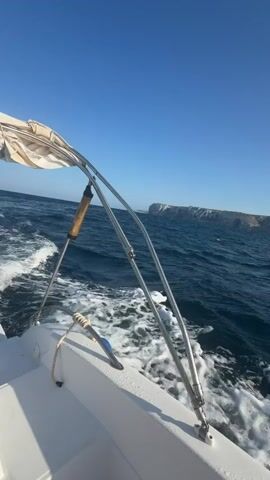 This video captures a journey on a boat in the ocean. The camera is positioned on the boat, looking out towards the water and the distant coastline. The water is a deep blue, with white foam trailing behind the boat, indicating movement. The sky is a clear, bright blue, with the sun shining. In the distance, a rocky cliff face rises out of the sea, and further beyond, hills or mountains are visible on the horizon. The boat itself appears to be white, with a metal railing and a fabric canopy visible in the foreground. The overall impression is one of a peaceful and scenic boat trip on a sunny day.