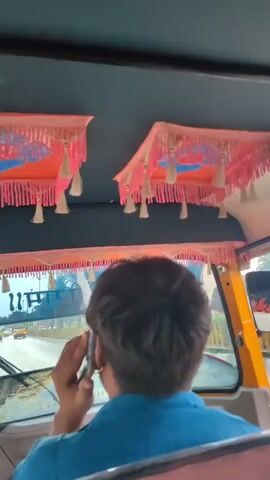 The video, taken from inside a vehicle, shows the back of a person's head as they hold a mobile phone to their ear and speak. The person is wearing a blue shirt. In front of them, through the windshield, a road with other vehicles can be seen. The interior of the vehicle features decorative pink and orange fabric with tassels hanging from the ceiling. The windshield has some text and images on it. The video is short and appears to capture a moment while traveling.