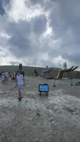 The video shows an outdoor scene with a group of people and several off-road vehicles. The ground is unpaved and appears to be a dirt or gravel path. In the background, there are rolling hills covered with vegetation, and the sky is partly cloudy.

In the foreground of the first frame, a young boy in a white t-shirt and grey shorts walks away from the camera. To his left, there is a large sculpture of a hand. A monitor is set up on a stand in the middle ground.

The second frame shows more people walking around a few vehicles. One of the vehicles is a dark SUV. There is also a small stall or kiosk in the middle ground with people gathered around it.

The third frame focuses on a group of people standing near several white off-road vehicles. A young boy in a teal t-shirt and dark shorts is in the foreground. Behind him, a woman in a bright yellow shirt and dark pants stands with another woman.

The fourth and fifth frames show a closer view of the off-road vehicles parked on the uneven terrain. The vehicles are robust and appear to be designed for off-road use. Some have modifications such as larger tires. The landscape continues to be hilly and natural.