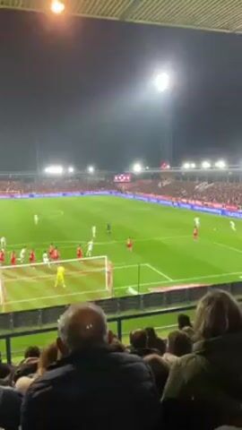 The video shows a football game being played in a stadium at night. The field is well-lit by stadium lights. There are two teams of players, one in red uniforms and the other in white. The game is in progress, with players moving around the field. In the foreground, there are spectators in the stands, including a man with a bald spot on the back of his head. The stadium is full of people. There is a scoreboard in the background, but the score is not clearly visible.