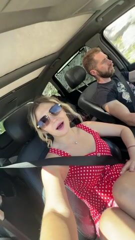 Two people, a woman and a man, are in the front seats of a car. The woman is in the passenger seat, and the man is driving. The woman is wearing a red and white polka dot dress and sunglasses. She is looking at the camera and appears to be talking or singing. The man is looking ahead at the road. The car is moving, as indicated by the blurred scenery outside the windows. The interior of the car is visible, including the dashboard and seats. The lighting suggests it is daytime.