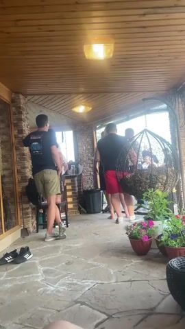 The video shows a group of people standing on a stone patio covered by a wooden ceiling with two lights. A man in a black t-shirt and khaki shorts is facing away from the camera, leaning against a chair and gesturing with his left hand. To his right, a group of men are standing together, one of whom is wearing red shorts. A large, black, hanging egg chair filled with cushions is visible on the right side of the frame, with potted flowers in front of it. The background shows a building with windows and a door. The floor is paved with large, irregular stones.