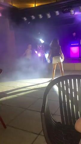 This video captures a lively scene at an outdoor venue at night. The environment appears to be a patio or open-air space adjacent to a multi-story building adorned with numerous lit windows, suggesting a hotel or resort. The focus is on a stage area, illuminated by dynamic purple and blue lights, with visible smoke effects enhancing the atmosphere. A disco ball hangs from the ceiling, reflecting beams of blue light across the floor and creating a party-like ambiance. Several individuals are present on and around the stage. One person, a male wearing a light-colored shirt and dark shorts, is seen dancing. Further back, two other individuals, possibly a female with dark hair in a white top and dark bottom, and another person are also visible on or near the stage area. The foreground of the video shows parts of two chairs, indicating seating for an audience or patrons. The overall impression is that of a nighttime event with music and entertainment.