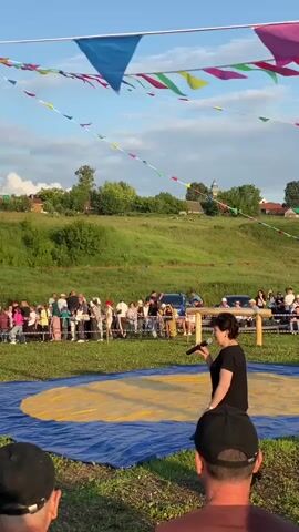 A woman in a black outfit holds a microphone and stands in front of a wrestling mat. A crowd of people watches from behind a fence, with a grassy hill in the background. Colorful triangular flags are strung across the sky. A Russian flag flies from a tall pole. A man in a suit walks toward the wrestling mat from the left side of the frame. The sun is setting, casting a warm glow on the scene. The overall atmosphere appears to be festive, suggesting an outdoor event or competition.