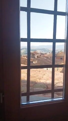 The video shows a view from a window with a barred pane, looking out over a picturesque Italian town. In the foreground, we see terracotta roof tiles, some appearing weathered and ancient. Beyond these, a cluster of buildings with similar terracotta roofs fills the middle ground, with glimpses of walls and windows suggesting a dense urban fabric. The background opens up to a rolling landscape of hills under a clear, bright sky. The light suggests it is daytime, likely late morning or early afternoon, with the sun casting a gentle glow on the scene. There are no people visible, but the presence of the buildings and rooftops implies a inhabited location. The barred window suggests a historical building, possibly a fortification or a prison, adding a layer of intrigue to the serene landscape outside. The overall impression is one of timeless beauty and a tranquil, sunny day in a charming Italian setting.