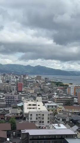 The video shows an overview of a densely populated city under a cloudy sky. The cityscape includes a mix of low-rise residential buildings and taller apartment complexes. In the background, a mountain range is visible. A body of water appears towards the horizon, possibly a bay or lake. The wind seems strong, evident from the audio. There are trains seen moving in the periphery.