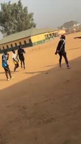 The video shows a group of young men playing soccer on a dirt field. There are several players wearing different colored shirts, including black, white, and red. The field is bordered by a building with a green and yellow facade. The sky appears hazy, and there are some trees visible in the background. The players are actively running and kicking the ball, with the camera panning to follow the action.
