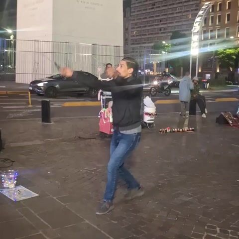 The video shows a man dancing in a public plaza at night. He is wearing a black jacket, blue jeans, and grey sneakers. He is dancing to music playing from a blue-lit speaker system nearby. In the background, there is a tall white monument with text on it, and several buildings. There are also other people present, including a man pushing a small cart with merchandise, some pedestrians, and a young girl. Cars are driving by on the street next to the plaza. The plaza is paved with bricks. There is a light-up display on the ground near the speaker system. The dancer gestures with his hands and moves around energetically.