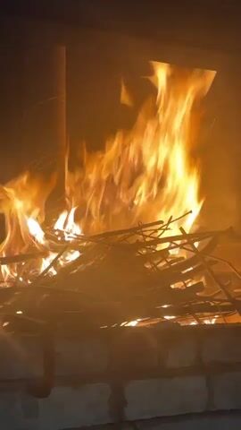 A close-up, static shot shows a fire burning in a brick hearth. The flames are vibrant orange and yellow, consuming a pile of dry sticks and branches. To the left, a metal pipe or chimney is visible. The background is dark, suggesting an indoor setting.