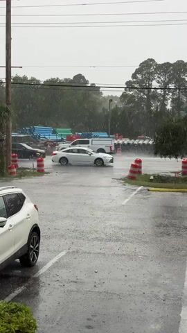 The video shows a heavy downpour in what appears to be a parking lot. Cars drive slowly through the rain, with water splashing from their tires. There are rows of pipes visible in the background, along with orange and white traffic cones. A sign for "Gulf Daddy's Seafood" is also visible. The parking lot is shiny with rainwater, and the sky is overcast.