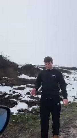 A young man stands in a snowy environment, wearing a black sweatshirt with a logo, black sweatpants, and brown boots. He is gesturing with his hands, apparently giving instructions to someone behind the camera, possibly to back up a car.  Snow covers the ground and a muddy embankment is visible in the background. The side mirror of a car is visible in the lower left corner of the frame. The sky is overcast and white.