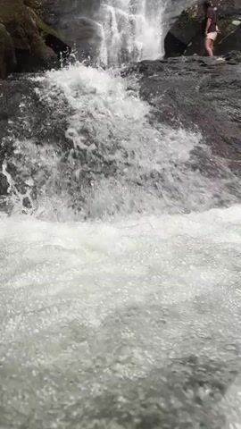 The video shows a waterfall cascading over rocks into a pool of water.  The water is white and foamy where it falls, and calmer in the pool below.  Two people are standing on the rocks beside the waterfall, appearing to be admiring the view.  The surrounding environment is lush with green foliage.