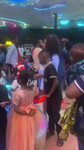 The video shows a crowd of people, mostly children and some adults, standing indoors. A young boy in a black polo shirt with "LONDON" printed on it stands near a woman with curly hair who is wearing a blue and white striped shirt over a white top. The boy looks down and to the side. A girl in a pink dress stands with her back to the camera. Other people are visible in the background, including a man in a blue jacket and a woman in a hijab. The environment appears to be a party or event space with bright lighting and decorations.