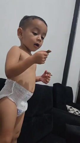 A young boy wearing a diaper stands on the right side of the frame, facing right and looking off-camera. He holds two small, brown objects in his hands. The boy's mouth is slightly open, and his gaze is directed away from the camera. Behind him, a dark sofa is visible, with a portion of a patterned cushion on its right side. The background is a plain, light-colored wall with a black vertical line running down the right edge. The lighting suggests an indoor setting. The video captures the boy's subtle movements and facial expression as he holds the objects.