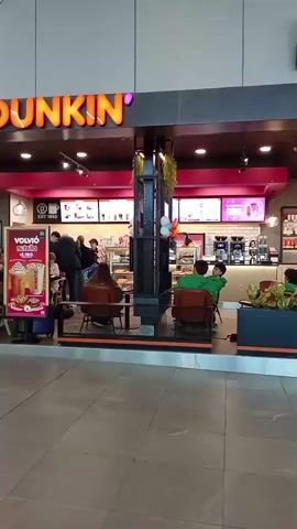 The video shows the exterior of a Dunkin' Donuts store. A sign with the Dunkin' logo is visible above the entrance. Inside the store, employees can be seen behind the counter, and customers are seated at tables. There are promotional posters displayed outside the store, one of which advertises a drink and has the price listed as 4.990. People are entering and exiting the store, and some are waiting in line. The overall atmosphere appears to be busy and normal for a retail establishment.