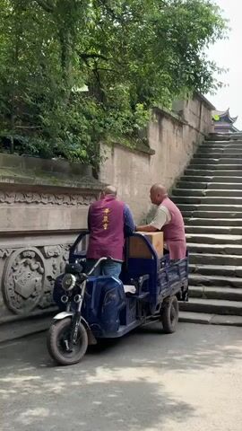 Two monks stand beside a blue tricycle on a street next to a flight of stairs. One monk is sitting in the back of the tricycle with a box. The other monk stands outside of the vehicle, facing him and the box. The standing monk wears a dark magenta vest, blue shirt, and blue jeans. The sitting monk also wears a magenta vest, with light beige pants. The vest of the standing monk has white script on the back. The stone wall behind the tricycle has ornate carvings in the wall.  A tree is growing behind the stone wall, and the staircase leads up behind the wall.