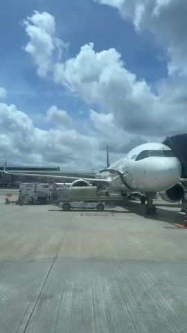 The video shows an airplane parked at an airport gate, connected to a jet bridge. The sky is blue with scattered white clouds. Several airport vehicles are visible around the plane, including a baggage tug and a bus. The camera pans across the scene, offering different perspectives of the aircraft and its surroundings.