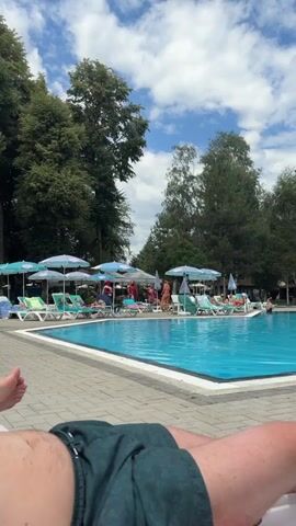 A swimming pool is visible, with lounge chairs and umbrellas around it. On the left, a person's legs are visible, lying on a lounge chair. In the background, two individuals in superhero costumes, one as Spiderman and the other as Iron Man, are standing near a group of people. Some of the people are sitting on lounge chairs, and others are standing. The environment appears to be outdoors, with trees and a cloudy sky visible.