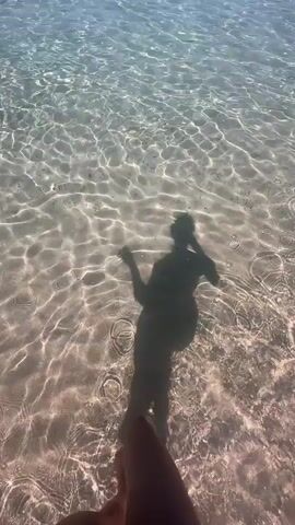 The video begins with a close-up, low-angle shot of a person's legs and feet submerged in crystal clear water. Sunlight refracts through the water, creating shimmering patterns on the sandy bottom. A shadow of the person taking the video, holding a phone, is visible in the water. The perspective shifts to show the person walking into the shallow water. The camera then pans up to reveal a wider view of a beach. The ocean is calm and clear, with visible sandy patches beneath the surface. In the distance, people are swimming and playing in the water. Several boats and inflatable water toys are also present near the shore. The sky is a clear, bright blue. The video ends with the camera still focused on the water and the sandy seabed.