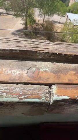 The video shows a close-up view of weathered wooden planks, possibly a railing or window frame, with peeling paint. In the background, an outdoor scene is visible, including trees, buildings, and a concrete ground area. The lighting suggests it is daytime, and the sky is clear. The camera moves slightly, offering a broader view of the outdoor environment. There are no people or specific actions clearly depicted, focusing instead on the textures of the wood and the general outdoor setting.