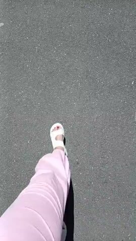 A first-person perspective video shows someone walking on an asphalt surface. The individual is wearing white sandals and pink pants. The camera angle is looking down at the person's feet and legs as they walk forward. The environment appears to be outdoors, with the asphalt suggesting a path, road, or sidewalk. The lighting suggests a sunny day.