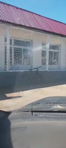 The video shows a slow panning shot from inside a vehicle, looking out at a building with a red roof. A bicycle is parked in front of the building, and there are bars on the windows. The video continues to pan, revealing more of the building and surrounding trees. A black car is parked in front of the building as well. The shot ends by panning upwards, showing more of the trees and a bright blue sky.