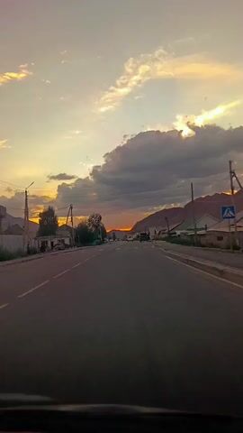 The video shows a view from a vehicle driving down a paved road. The road has white dashed lines and a pedestrian crossing with yellow and white stripes. On the left side of the road, there are houses, trees, and a utility pole with wires. On the right side, there are more houses and a mountain range in the distance. The sky is filled with clouds and the sun is setting, casting an orange glow. There are a few cars visible on the road.