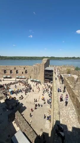 From an elevated position overlooking a courtyard within a large stone structure, a scene unfolds on a sunny day under a clear blue sky with a few wispy clouds. A river, bordered by trees, is visible beyond the structure. The courtyard is filled with people, many of whom are gathered around a central area. There, four individuals dressed in medieval attire engage in a staged fight. Two appear to be armed with spears, while the other two wield axes. Surrounding them, spectators stand behind a wooden barrier, observing the spectacle. Scattered throughout the courtyard are several white-and-blue striped tents or stalls, likely part of a market or fair. The stone structure itself is characterized by its aged appearance, with weathered walls and arched openings. The ground of the courtyard is paved with stone, enhancing the historical atmosphere of the scene.