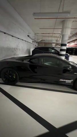 The video shows a black McLaren sports car parked in an underground parking garage. It is parked in a designated space with painted white lines, and there is another blue car parked behind it. The garage has a concrete ceiling with visible pipes and lighting fixtures. The support columns are painted with black and white stripes.