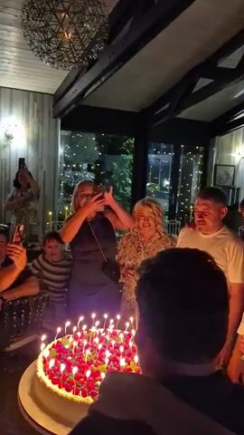 In this video, a group of people are gathered around a large birthday cake adorned with raspberries and lit candles. The scene appears to be an indoor celebration, possibly a restaurant or private party, with elegant decor including a modern chandelier and decorative lighting visible through a large window. The time of day is evening, suggested by the indoor lighting and the dark exterior visible through the window, which shows twinkling lights that could be from a town or an outdoor event.

The central activity is the singing of "Happy Birthday" to someone whose face is obscured from the camera's view by the cake. A woman in a dark dress is in the foreground, holding up a phone to record the moment and clapping her hands. Other individuals, including men, women, and a child, are leaning in towards the cake, their faces reflecting the candlelight. Their expressions suggest joy and participation in the celebration. The overall mood is festive and heartwarming.

The individuals are dressed in casual to semi-formal attire. The cake itself is a prominent object, decorated with fresh fruit and numerous small candles, all of which are alight. The setting features wooden beams on the ceiling and paneled walls, contributing to a warm and inviting atmosphere. There is no visible text in the video. The location context of Zelyonovka, Russia, places this intimate birthday celebration in a specific geographical setting.